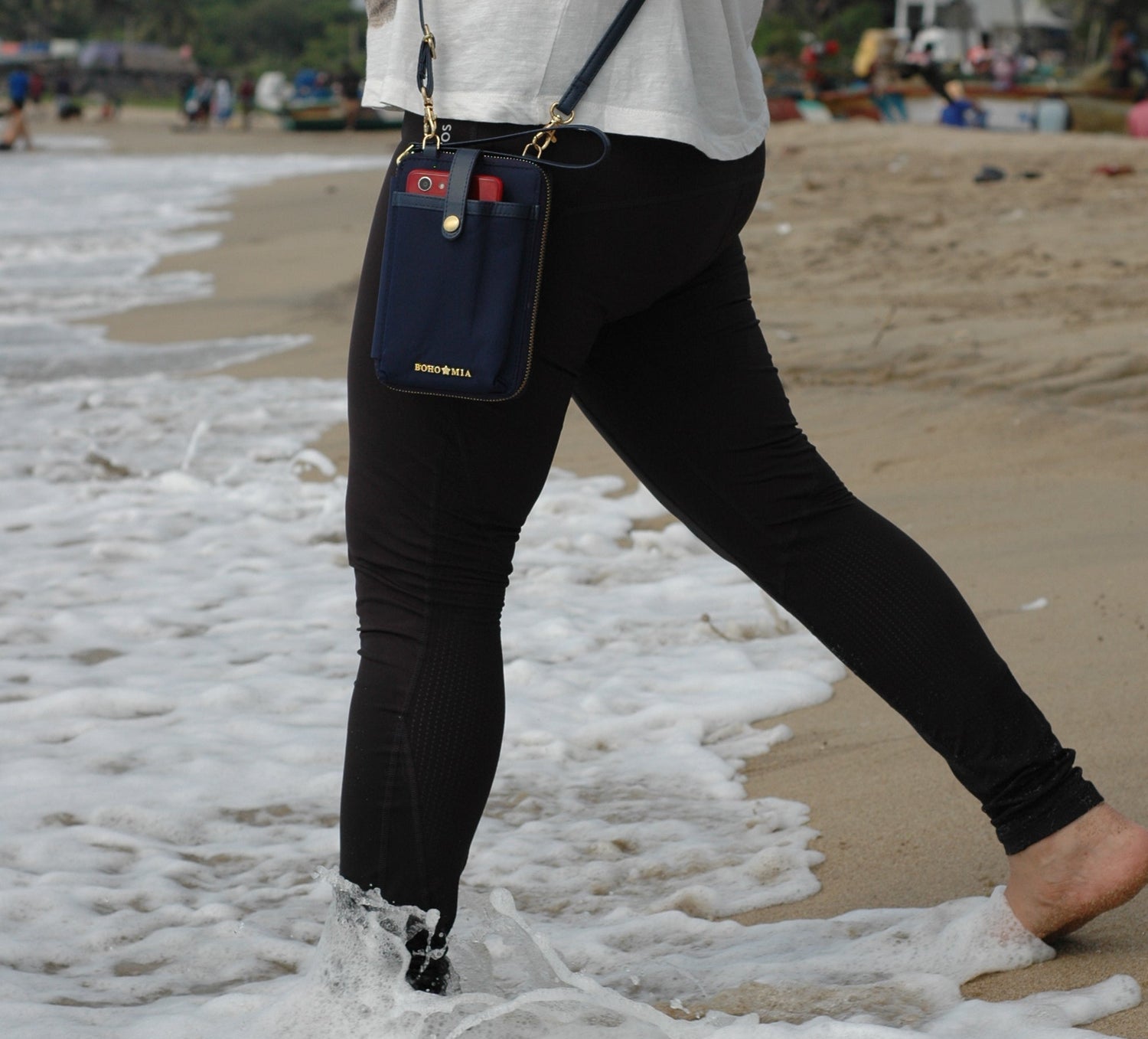 Person walking on a beach with a blue breeze mobile sling wallet with waves crashing on her feet